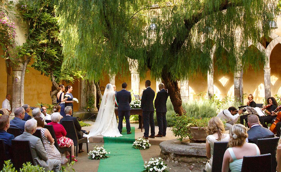 Bride and groom stand at the alter facing away from the camera. The aisle has a green cloth down the center with white flower bouquets on each side. Sitting on each side of the aisle are guests. Behind the bride and groom is a white and yellow building with large arches. They stand under a large drooping green tree.